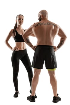 Handsome bald, tattooed fellow in black shorts and sneakers, standing back, with attractive brunette lady in leggings and top are posing isolated on white background and looking at the camera. Fitness couple, chic muscular bodies, gym concept. The love story.