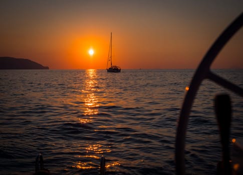 The sun is setting over the ocean with a sailboat in the distance and a helm in the foreground. Photo of a colorful sunset over the ocean with a sailboat in the distance