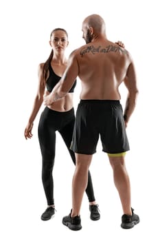 Good-looking bald, tattooed man in black shorts and sneakers, standing back, with alluring brunette woman in leggings and top are posing isolated on white background and looking at the camera. Fitness couple, chic muscular bodies, gym concept. The love story.