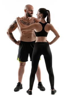 Handsome bald, tattooed guy in black shorts and sneakers with attractive brunette girl in leggings and top, standing back, are posing isolated on white background and looking at each other. Fitness couple, chic muscular bodies, gym concept. The love story.