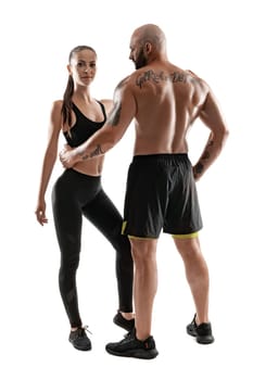 Good-looking bald, tattooed male in black shorts and sneakers with alluring brunette female in leggings and top are posing standing sideways isolated on white background and looking at the camera. Fitness couple, chic muscular bodies, gym concept. The love story.
