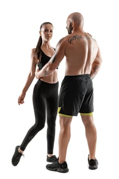 Good-looking bald, tattooed fellow in black shorts and sneakers, standing back, with alluring brunette lady in leggings and top are posing isolated on white background and looking at the camera. Fitness couple, chic muscular bodies, gym concept. The love story.