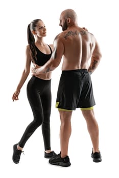 Stately bald, tattooed man in black shorts and sneakers with pretty brunette woman in leggings and top are posing standing sideways isolated on white background and looking at each other. Fitness couple, chic muscular bodies, gym concept. The love story.