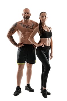 Athletic bald, tattooed man in black shorts and sneakers with cute brunette woman in leggings and top are posing isolated on white background and looking at the camera. Fitness couple, chic muscular bodies, gym concept. The love story.