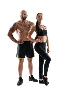 Athletic bald, tattooed male in black shorts and sneakers with cute brunette female in leggings and top are posing isolated on white background and looking at the camera. Fitness couple, chic muscular bodies, gym concept. The love story.