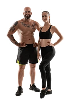 Athletic bald, tattooed fellow in black shorts and sneakers with cute brunette lady in leggings and top are posing isolated on white background and looking at the camera. Fitness couple, chic muscular bodies, gym concept. The love story.