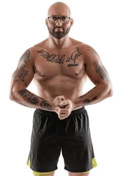 Athletic bald, bearded, tattooed man in glasses, black shorts is posing and showing his muscles isolated on white background, looking at the camera. Chic muscular body, fitness, gym, healthy lifestyle concept. Close-up portrait.