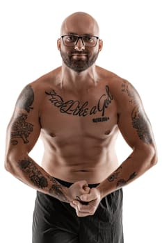 Athletic bald, bearded, tattooed male in glasses, black shorts is posing and showing his muscles isolated on white background, looking at the camera. Chic muscular body, fitness, gym, healthy lifestyle concept. Close-up portrait.