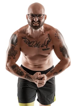 Athletic bald, bearded, tattooed guy in glasses, black shorts is posing and showing his muscles isolated on white background, looking at the camera. Chic muscular body, fitness, gym, healthy lifestyle concept. Close-up portrait.
