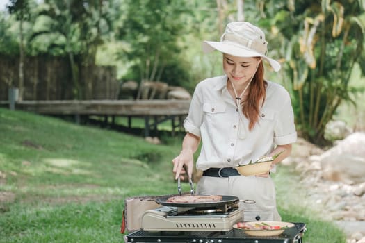 Beautiful Asian woman in her 30s preparing food Grill pork chop steak for dinner on the picnic grill. Adventure camping vacation ideas.