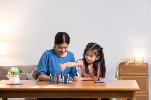 A young family spends their free time together in the living room at home. Mother and little daughter draw pictures with crayons on paper, smiling happy..