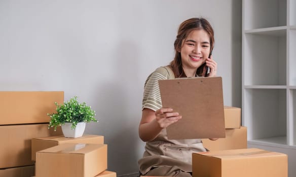 Small business entrepreneur SME freelance woman using phone call receive from customer checking product on stock at home office, online marketing packaging delivery box, SME e-commerce concept..