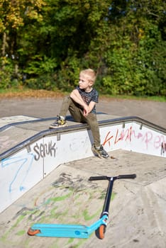 Boy on scooter makes a trick and enjoying his riding in the skate park at cloudy spring day. Young man doing trick on the kick scooter in the park. Full body little boy on the kick scooter on sunny autumn day in skate park. From below of active teen boy with kick scooter standing on ramp in skate park while preparing for performing trick in sunny spring day.