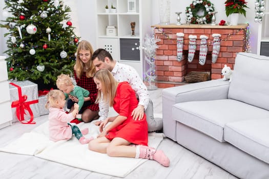 Two women, a man and two children spend Christmas together. The family sits on a white carpet in front of the fireplace. The fireplace is decorated with colorful lights. Presents lie under the large Christmas tree.