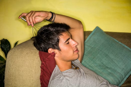 A man laying on a couch with a head massager in his hand.Photo of a young and handsome man relaxing on a couch with a head massager in hand