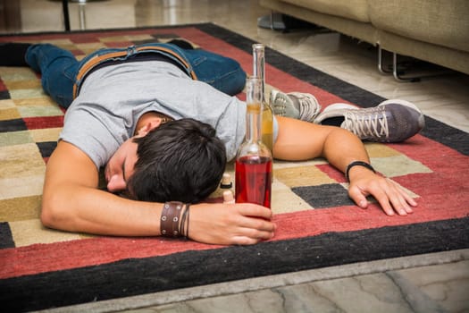 A man laying on the floor next to a bottle of alcohol. Photo of a young and handsome man lying on the floor next to a bottle of alcohol