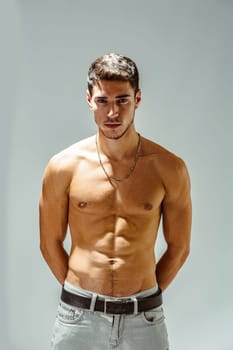 A shirtless handsome young man posing for a picture showing muscular torso, looking at camera, wearing grey jeans