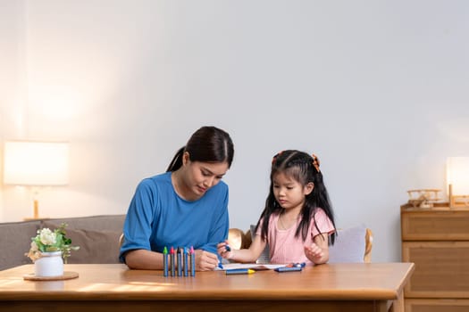 A young family spends their free time together in the living room at home. Mother and little daughter draw pictures with crayons on paper, smiling happy..