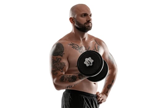 Good-looking bald, bearded, tattooed guy in black shorts is posing holding a dumbbell in his hand and looking away, isolated on white background . Chic muscular body, fitness, gym, healthy lifestyle concept. Close-up portrait.