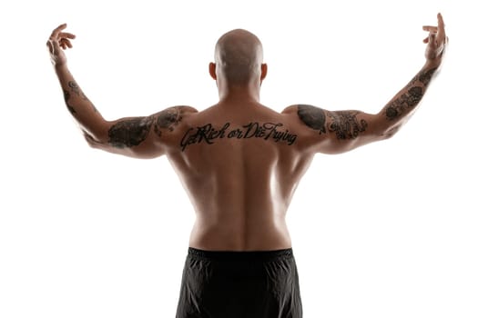 Stately bald, bearded, tattooed male in black shorts is posing standing back to the camera, raised his hands and showing muscles, isolated on white background. Chic muscular body, fitness, gym, healthy lifestyle concept. Close-up portrait.