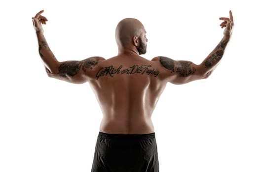 Stately bald, bearded, tattooed guy in black shorts is posing standing back to the camera, raised his hands and showing muscles, isolated on white background. Chic muscular body, fitness, gym, healthy lifestyle concept. Close-up portrait.