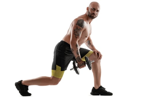 Stately bald, bearded, tattooed person in black shorts and sneakers is posing with a dumbbell in his hand, standing sideways and looking at the camera, isolated on white background. Chic muscular body, fitness, gym, healthy lifestyle concept. Full length portrait.