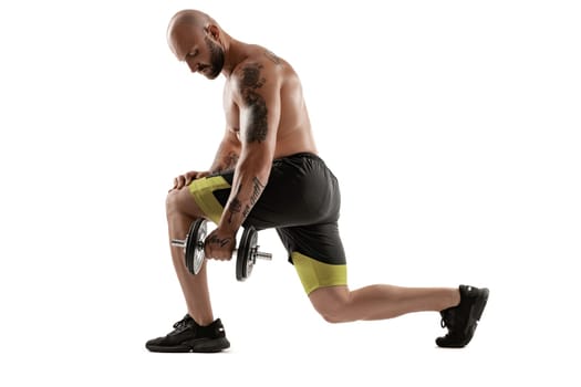 Strong bald, bearded, tattooed man in black shorts and sneakers is posing with a dumbbell in his hand, standing sideways isolated on white background. Chic muscular body, fitness, gym, healthy lifestyle concept. Full length portrait.