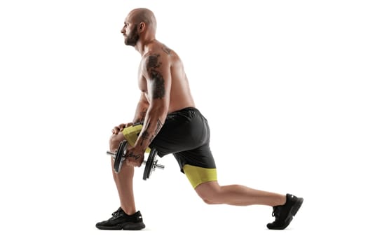 Strong bald, bearded, tattooed guy in black shorts and sneakers is posing with a dumbbell in his hand, standing sideways isolated on white background. Chic muscular body, fitness, gym, healthy lifestyle concept. Full length portrait.