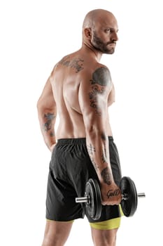 Masculine bald, bearded, tattooed male in black shorts is posing standing sideways, with a dumbbell in his hand, isolated on white background. Chic muscular body, fitness, gym, healthy lifestyle concept. Close-up portrait.