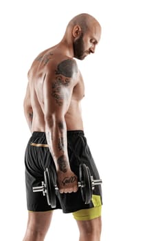 Masculine bald, bearded, tattooed guy in black shorts is posing standing sideways, with a dumbbell in his hand, isolated on white background. Chic muscular body, fitness, gym, healthy lifestyle concept. Close-up portrait.