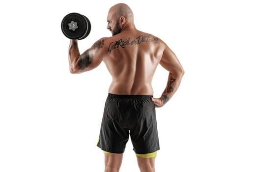 Masculine bald, bearded, tattooed person in black shorts is posing with a dumbbell in his hand, standing back, isolated on white background. Chic muscular body, fitness, gym, healthy lifestyle concept. Close-up portrait.