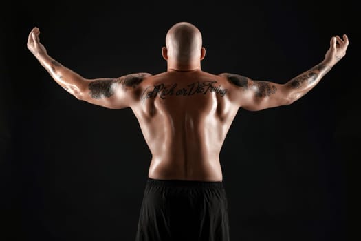 Athletic bald, bearded, tattooed male in black shorts is posing standing back to the camera and showing his muscles, standing against a black background. Chic muscular body, fitness, gym, healthy lifestyle concept. Close-up portrait.