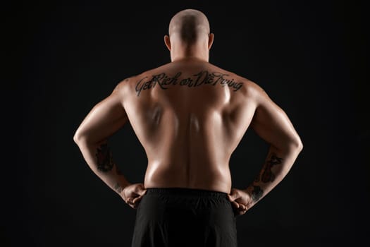 Athletic bald, bearded, tattooed guy in black shorts is posing standing back to the camera and showing his muscles, standing against a black background. Chic muscular body, fitness, gym, healthy lifestyle concept. Close-up portrait.
