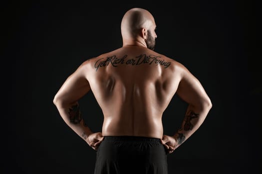 Athletic bald, bearded, tattooed fellow in black shorts is posing standing back to the camera and showing his muscles, standing against a black background. Chic muscular body, fitness, gym, healthy lifestyle concept. Close-up portrait.