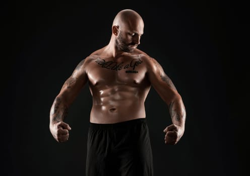 Athletic bald, bearded, tattooed person in black shorts is demonstating his muscles posing against a black background and looking down. Chic muscular body, fitness, gym, healthy lifestyle concept. Close-up portrait.