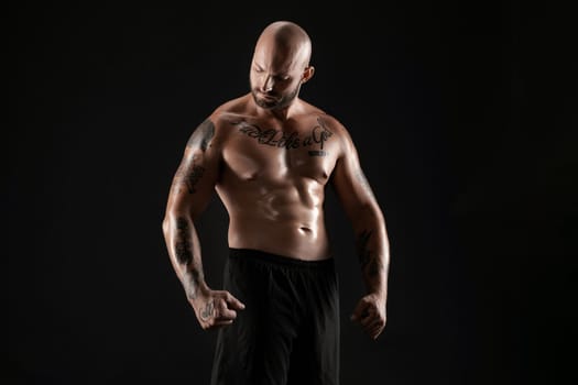 Handsome bald, bearded, tattooed man in black shorts is demonstating his muscles and looking at it while posing against a black background. Chic muscular body, fitness, gym, healthy lifestyle concept. Close-up portrait.