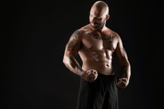 Handsome bald, bearded, tattooed guy in black shorts is demonstating his muscles and looking at it while posing against a black background. Chic muscular body, fitness, gym, healthy lifestyle concept. Close-up portrait.