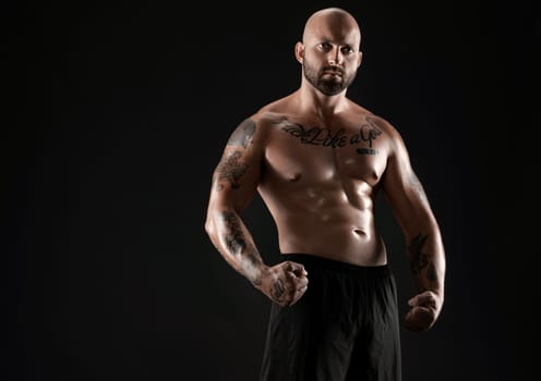 Handsome bald, bearded, tattooed male in black shorts is demonstating his muscles posing against a black background and looking at the camera. Chic muscular body, fitness, gym, healthy lifestyle concept. Close-up portrait.