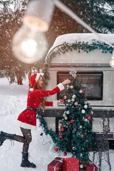 Young woman in santa costume decorates the Christmas tree at winter campsite getting ready for the new year. New year celebration concept