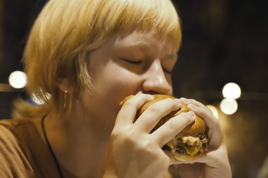 A teenager girl eats a hamburger in a restaurant, enjoying a delicious juicy burger. Close-up.