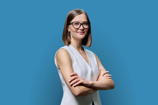 Young smiling woman in glasses with crossed arms posing on blue studio background. Positive business confident successful female looking at camera with folded hands. Business, work, training, people