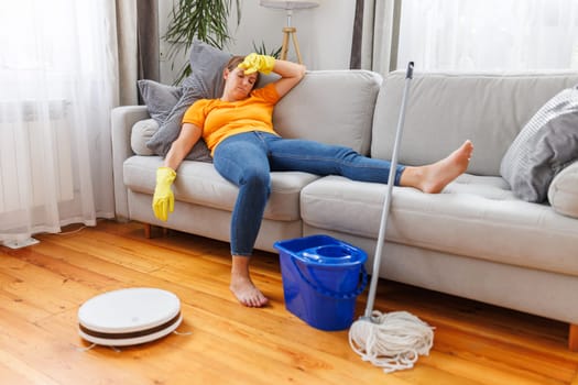 Robotic vacuum cleaner cleaning a room while tired woman relaxing on the sofa after doing housework. Concept of the advantages of modern cleaning technologies, buying a robot vacuum cleaner