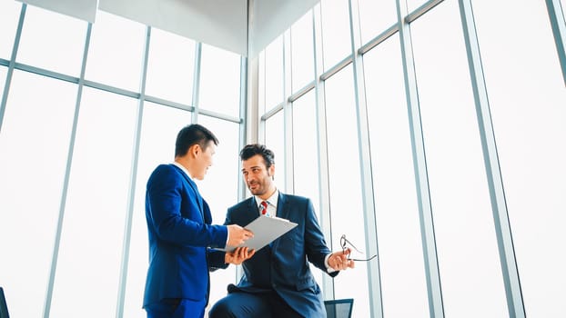 Two business people talk project strategy at office meeting room. Businessman discuss project planning with colleague at modern workplace while having conversation and advice on financial report. Jivy