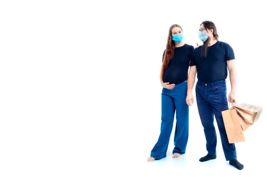young pregnant woman and her husband with shopping bags in medical masks. safety from colds flu and coronavirus on white background. Pregnancy shopping concept happy young family with shopping bags