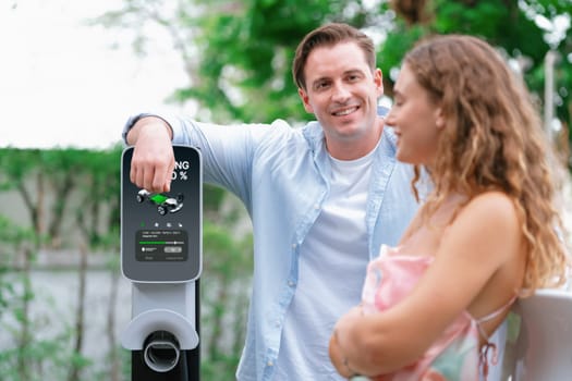 Happy and lovely couple with eco-friendly conscious recharging electric vehicle from EV charging station. EV car technology utilized as alternative transportation for future sustainability. Synchronos