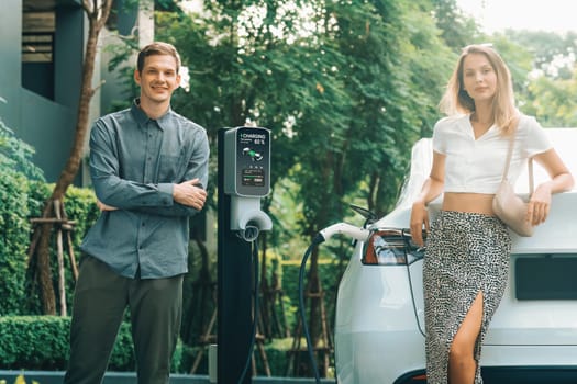 Young couple travel with EV electric car charging in green sustainable city outdoor garden in summer shows urban sustainability lifestyle by green clean rechargeable energy of electric vehicle innards