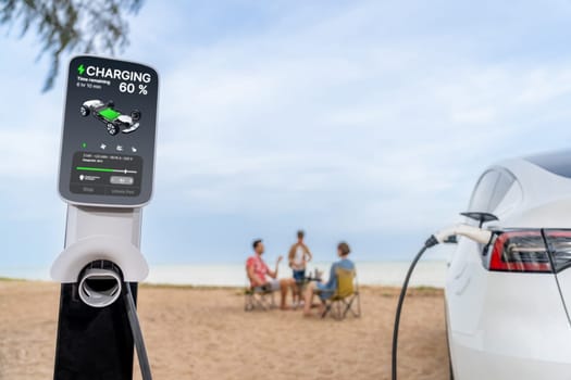 Alternative family vacation trip traveling by the beach with electric car recharging battery from EV charging station with blurred family enjoying the seascape background. Perpetual