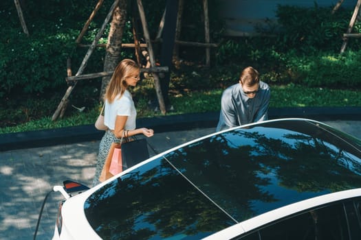 Young couple travel with EV electric car charging in green sustainable city outdoor garden in summer shows urban sustainability lifestyle by green clean rechargeable energy of electric vehicle innards