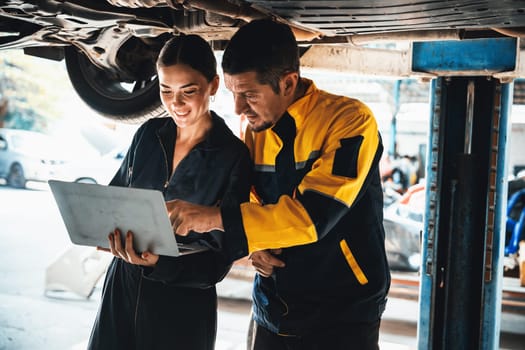 Two vehicle mechanic working together underneath lifted car, conduct car inspection with laptop. Automotive service technician in uniform carefully make diagnostic troubleshooting. Oxus