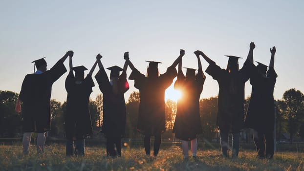 College graduates in robes holding hands at sunset